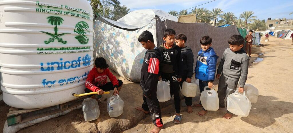 Children fill jerrycans with water from a large white UNICEF water tank in a shelter area in Gaza.